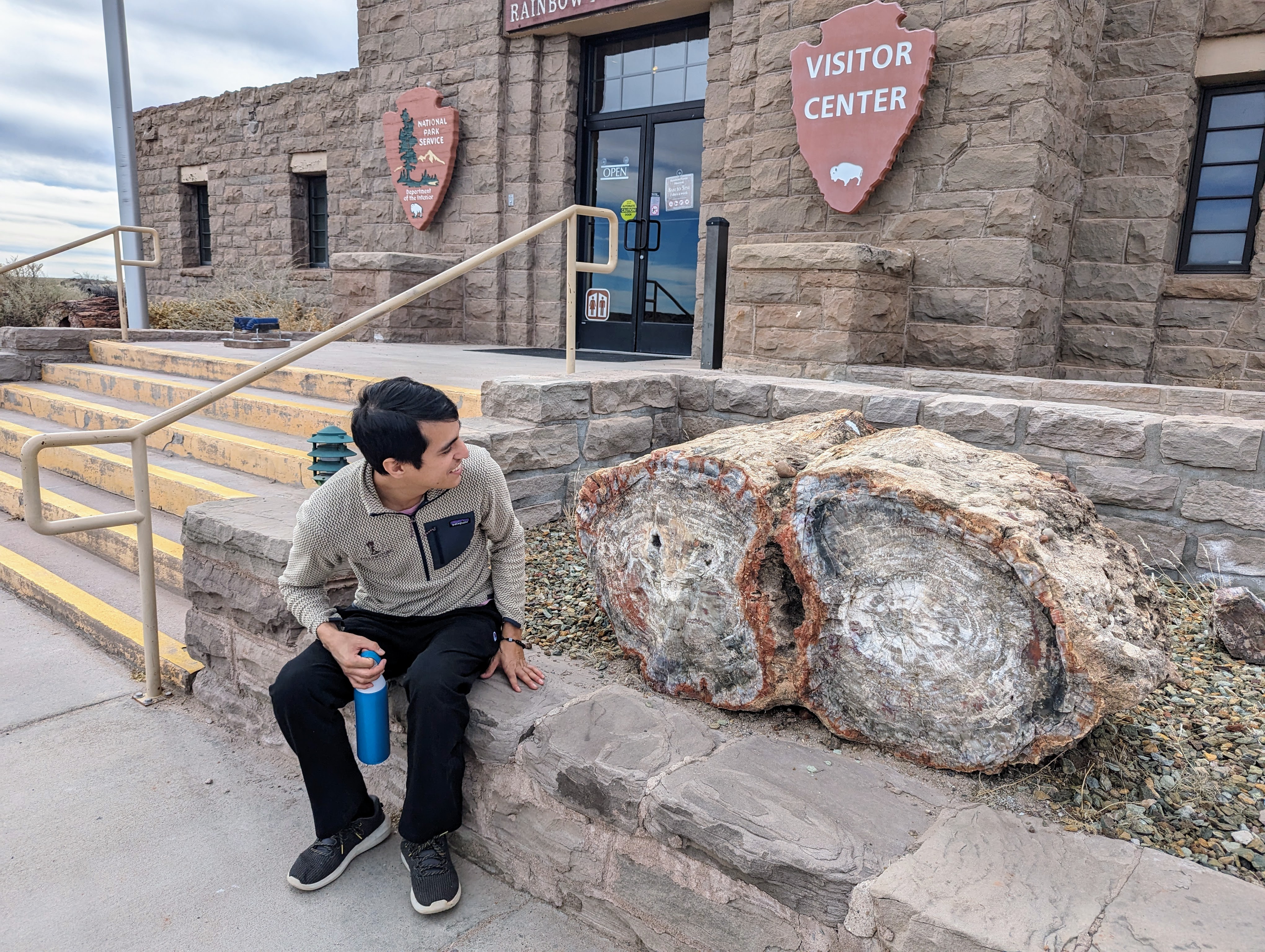 A visitor sitting beside a large, polished petrified log outside the Rainbow Forest Visitor Center at the south entrance of the park.